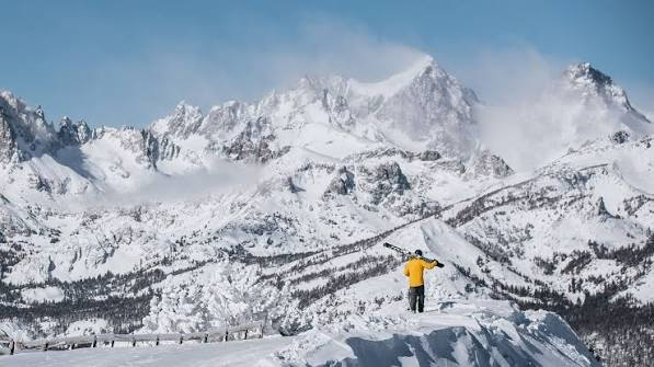 A skier in a yellow jacket standing on a summit, skis over one shoulder, jagged peaks behind