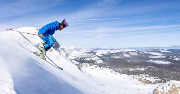 A skier in a blue jacket carving off the lip of a steep pitch above a wide valley