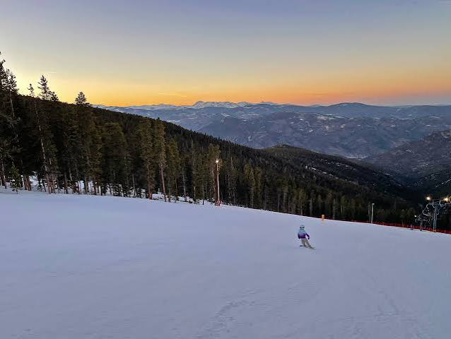 A lone skier on a wide groomed run at dusk with orange light over the Front Range
